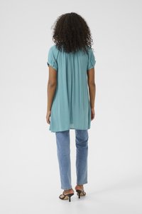 Tunique bleu clair à manches courtes, avec un dos plissé, associée à un jean en denim clair et des talons ouverts noirs. Fond uni.