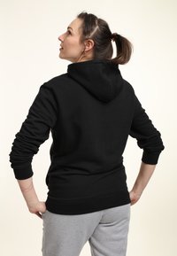 Felpa con cappuccio nera dal taglio rilassato, con polsini a coste e tasca a marsupio. Il soggetto è posato di schiena indossando pantaloni della tuta grigi.
