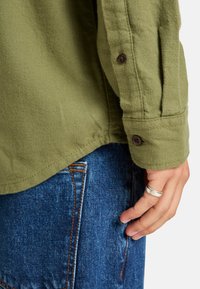 Camisa verde oliva con una tela texturizada, puños abotonados y un corte relajado, combinada con jeans de denim azul con costuras naranjas.