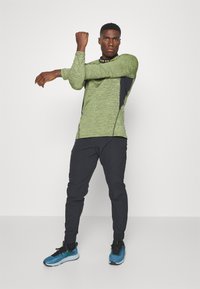 Lichtgroene lange mouw sportshirt met zwarte accenten, gecombineerd met zwarte tapered broek en blauwe sportschoenen. Beschikt over een ronde halslijn.