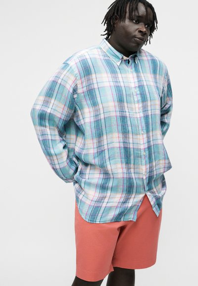 Osoba s krátkymi dredmi, oblečená v voľnej modro-zelenkavej kockovanej košeli na gombíky a koralových šortkách, stojaca s rukami za chrbtom.