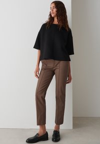 Maglietta nera oversize a maniche corte abbinata a pantaloni marroni a gamba dritta, con cuciture sottili e mocassini neri, su uno sfondo neutro.