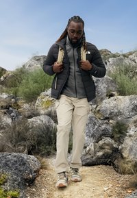 Casaco isolado preto sobre um fleece cinza com fecho, calças bege e sapatos de caminhada multicoloridos. O fundo apresenta um terreno rochoso e vegetação.