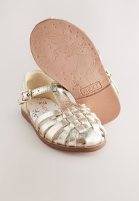 Goldene Metallsandalen mit gewebtem Riemen-Design, verstellbarer Schnalle und einem gemusterten Fußbett. Die Sohle zeigt ein Einhornmotiv.