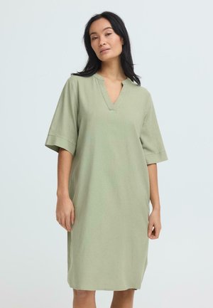 Kobieta o długich ciemnych włosach, ubrana w luźną, sięgającą kolan sukienkę w kolorze szałwiowym, z rękawami do łokci i dekoltem w kształcie litery V.