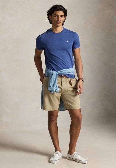 Férfi áll, kezét zsebre téve, kék pólót, bézs rövidnadrágot, világoskék inget deréknál megkötve és fehér tornacipőt viselve, semleges háttér előtt.