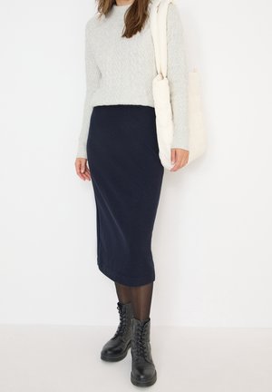 Frau in grauem Kabelstrickpullover, marineblauem Midirock, schwarzen Schnürstiefeln und weißer, gepolsterter Umhängetasche vor weißem Hintergrund.