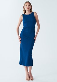 Abito midi aderente di colore blu con tessuto strutturato, scollo ampio e spacco laterale. Indossato con sandali con tacco alti di tonalità neutra.