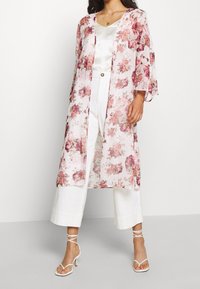 Žena v bílých širokých kalhotách a saténovém topu, přikrytá průhledným květinovým kimono a bílými sandály na podpatku.