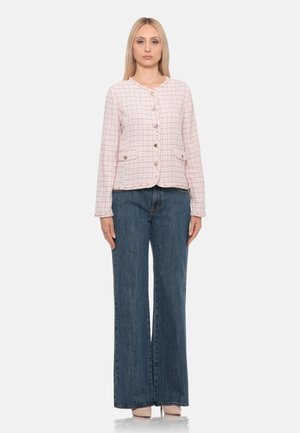 Donna che indossa una giacca rosa strutturata con bottoni e tasche frontali, jeans blu a vita alta e gamba larga, e scarpe beige, in piedi dritta.