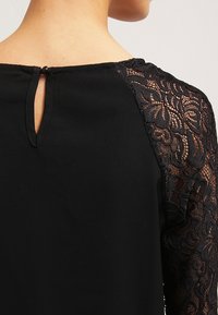 Blusa nera con maniche in pizzo con motivi floreali, apertura a forma di chiave sul retro e una consistenza del tessuto morbida e liscia.