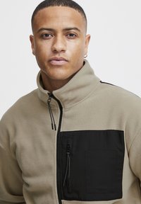 Chaqueta de forro polar en beige con bolsillo de tela negra. Cuello alto y cremallera frontal. Presenta dobles cremalleras en el bolsillo izquierdo y una textura suave.