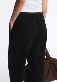 Persoană purtând pantaloni negri cu talie elastică și un top alb, purtând o geantă mare maro, cu mâna în buzunar, cu fața spre departe de cameră.