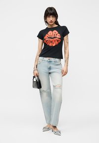 Γυναίκα που φορεί μαύρο t-shirt Marc Jacobs, ανοιχτόχρωμο τζιν με σκισίματα, ασημί μυτερές γόβες, κρατά μικρή μαύρη τσάντα, στέκεται μπροστά σε απλό φόντο.
