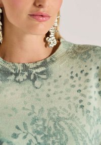 Close-up van een persoon die een groen gebreid truitje met patroon en grote hangende witte bloemenoorbellen draagt.