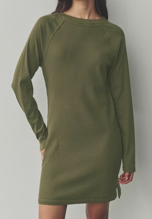 Femme portant une robe en tricot côtelé vert olive à manches longues, debout devant un fond clair uni.