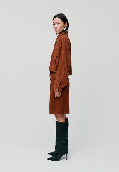 Braune Wildlederjacke und Rockensemble, das lange schwarze Stilettos umfasst. Glatte Texturen mit einem modernen, cropped Schnitt und Knopfakzenten.
