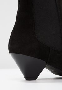 Bota de tobillo de ante negro con un panel elástico texturizado, que presenta un tacón en forma de cono con una base negra lisa. Vista en primer plano desde un lado.