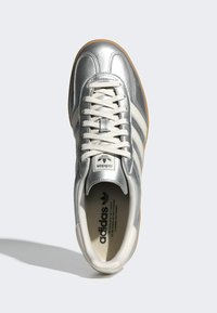 Silberne Leder-Sneaker mit drei weißen Streifen, runder Spitze und einer beige Gummisohle. Die Schnürsenkel sind hellgrau und das Branding befindet sich auf der Zunge.