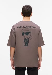 Man in een bruin oversized t-shirt met een cartoonafbeelding van Karl Lagerfeld in een pak en met zonnebril op de rug, staand tegen een witte achtergrond.
