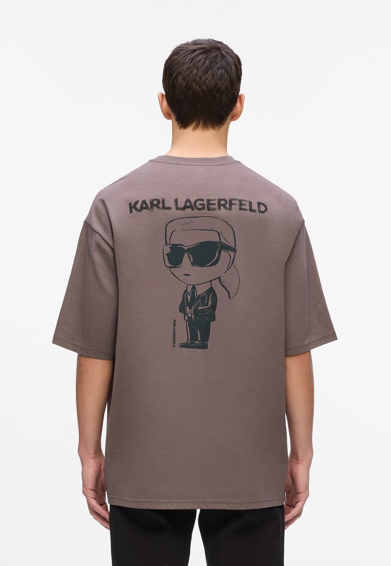 Man in een bruin oversized t-shirt met een cartoonafbeelding van Karl Lagerfeld in een pak en met zonnebril op de rug, staand tegen een witte achtergrond.