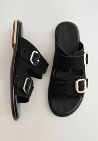 Sandales en suede noir avec deux sangles réglables et boucles en or, semelle rembourrée et semelle lisse. Design plat adapté à un usage décontracté.
