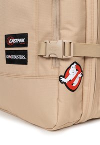 Beige Eastpak ryggsäck med en slät textur, med två svarta logotyper och ett röd-vitt Ghostbusters-emblem. Vadderade justerbara remmar.