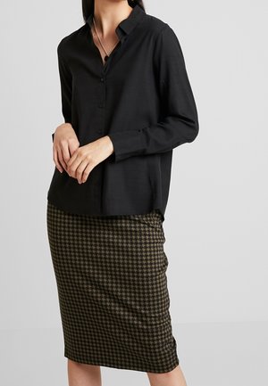 Kobieta ubrana w czarną koszulę na guziki i ołówkową spódnicę do kolan w brązowo-czarną pepitkę, stojąca na jednolitym tle.