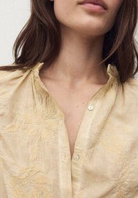 Donna con lunghi capelli castani che indossa una camicetta beige chiaro con bottoni, decorata con ricami floreali e un colletto arricciato.