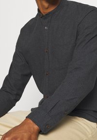 Hombre vestido con una camisa de botones gris oscuro y un cuello alto, y pantalones beige, sentado con los brazos relajados.