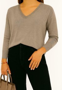 Beige v-hals trui met geribde manchetten, gedragen met zwarte jeans. De trui heeft een zachte textuur, een ontspannen pasvorm en een lichte drap.