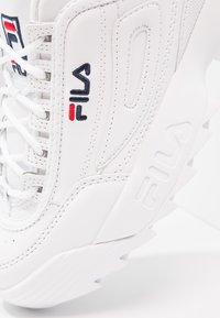 Zapatilla deportiva blanca con parte superior texturizada, que presenta un logo bordado en rojo y azul, suela de goma con elementos de tracción y cierre de cordones.