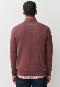 Bordeauxroter Strickpullover mit hohem Kragen, gerippten Bündchen und Saum. Verfügt über ein strukturiertes Muster und ist über einem rot gestreiften Hemd getragen.