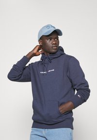 Sudadera azul marino con bolsillo frontal y cordón, con el logotipo en blanco "TOMMY JEANS". Gorra azul clara con "DAD" bordado en la parte frontal.