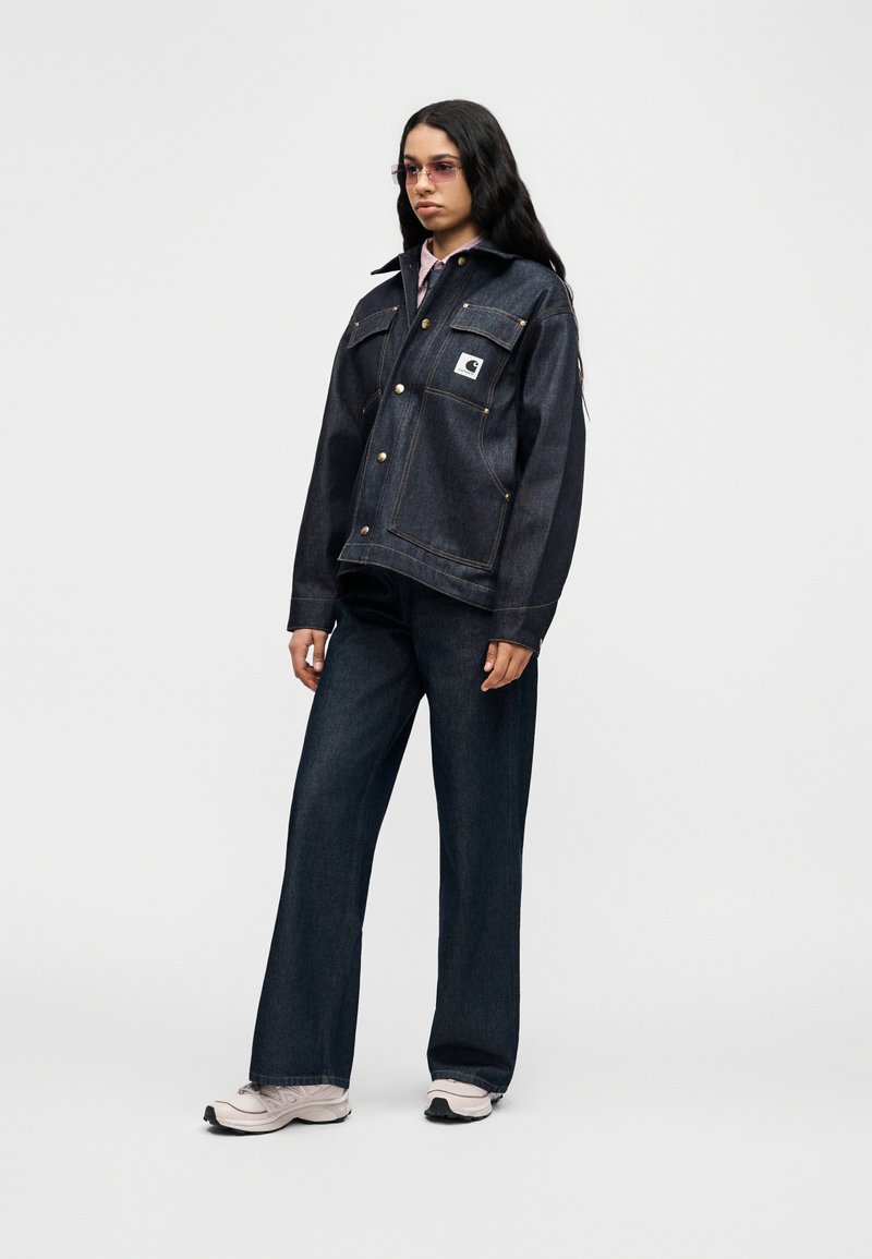 Veste en jean foncé avec fermeture à boutons et deux poches avant ; portée avec un jean large foncé et des baskets de couleur claire.