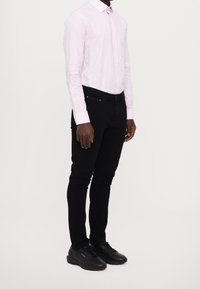 Chemise à rayures rose clair à manches longues, jean noir slim-fit et chaussures de sport noires, mettant l'accent sur un design simple et une silhouette ajustée.