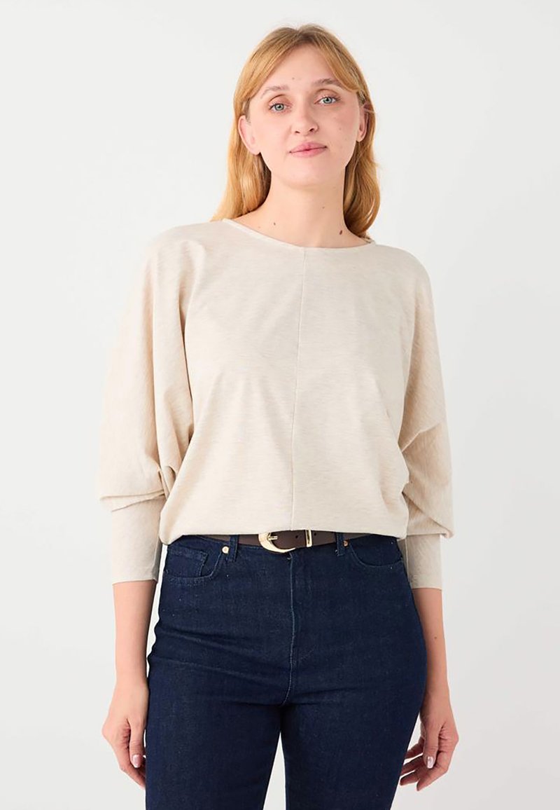 Femme blonde portant un haut beige à manches longues froncées et un jean taille haute bleu foncé avec une ceinture marron.