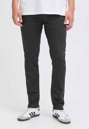 Modèle portant un pantalon noir à coupe ajustée, un T-shirt blanc et des baskets blanches à rayures noires sur un fond uni clair.