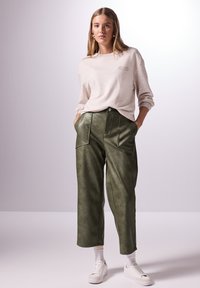 Ljusbeige sweatshirt med en subtil logotyp, kombinerad med mörkgröna byxor i konstläder med vida ben som har framfickor och knappstängning. Vita sneakers.