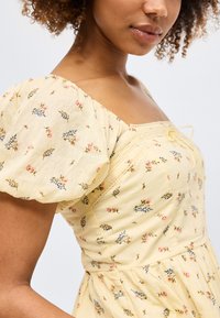 Γυναίκα που φοράει κίτρινο φόρεμα με φουσκωτά κοντά μανίκια και μικρό ροζ και μπλε λουλουδάτο σχέδιο σε λευκό φόντο.