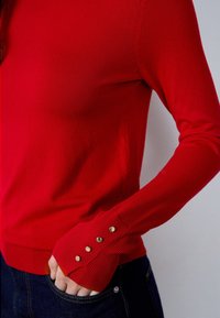 Red knit sweater with a fitted design, featuring ribbed cuffs and three gold buttons at the sleeve openings. Soft texture.