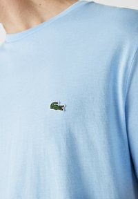 Camisa de algodón de color azul claro con un logo de cocodrilo bordado en verde en el lado izquierdo del pecho. Textura suave y escote clásico tipo crew.