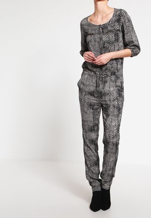 Nainen pukeutuneena mustavalkoiseen, kuvioituun, pitkähihainen haalariin, jossa on kiristysnauha vyötäröllä, ja mustiin nilkkureihin, seisomassa yksivärisen taustan edessä.