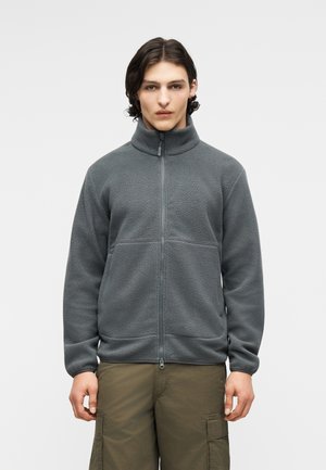 Νεαρό άτομο με μεσαίου μήκους σκούρα μαλλιά που φοράει γκρι φλις μπουφάν με φερμουάρ και χακί παντελόνι cargo, στέκεται μπροστά σε απλό φόντο.