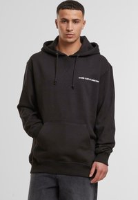 Black hooded sweatshirt with a kangaroo pocket, featuring white text "DJINNS CLUB DE L'ACTION" on the left chest. Made of textured fabric.