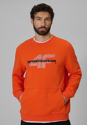 Mężczyzna z brodą, ubrany w pomarańczową bluzę z logo "4F SPORTSWEAR", ręce w przedniej kieszeni, neutralne szare tło.