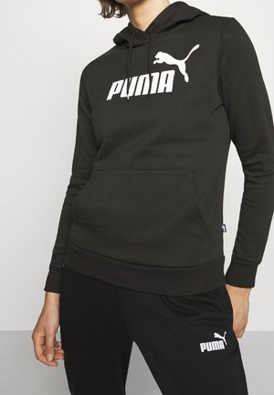 Osoba w czarnej bluzie z kapturem Puma i czarnych spodniach dresowych Puma, stoi z jedną ręką luźno u boku.