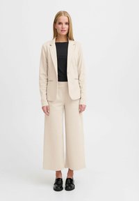 Beige blazer en wijde broek set, gecombineerd met een geribbeld zwart topje. Glad materiaal, getailleerde blazer, losse broek. Zwarte schoenen.