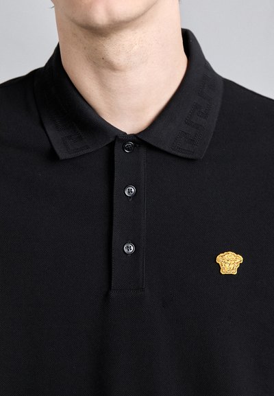 Polo noir avec un col texturé arborant un logo. Il possède trois boutons et un ornement doré proéminent sur la poitrine.