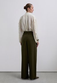 Mujer con el cabello recogido en un moño, vistiendo una blusa crema transparente con mangas acampanadas y pantalones de pierna ancha de talle alto en color oliva, de pie contra una pared lisa.